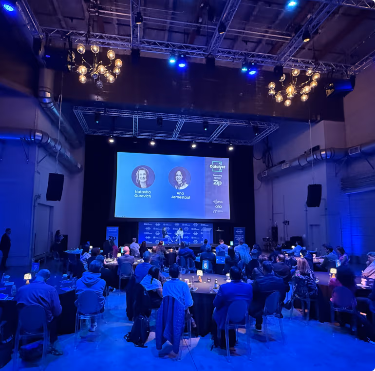 Attendees at round tables in a blue-lit hall, with two Catalyst session speakers on the big screen